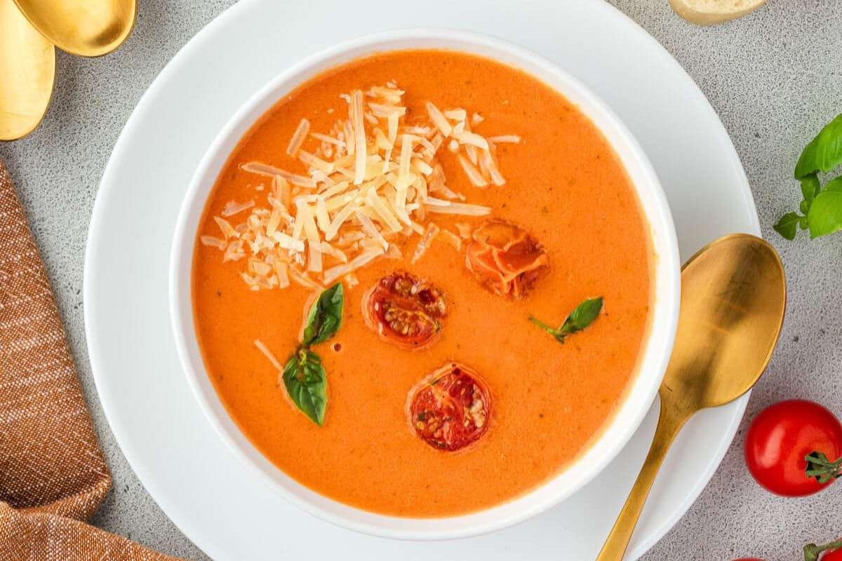 A bowl of creamy tomato soup, perfect for summer soups, topped with shredded cheese, roasted cherry tomatoes, and basil leaves, served with a gold spoon on a white plate.