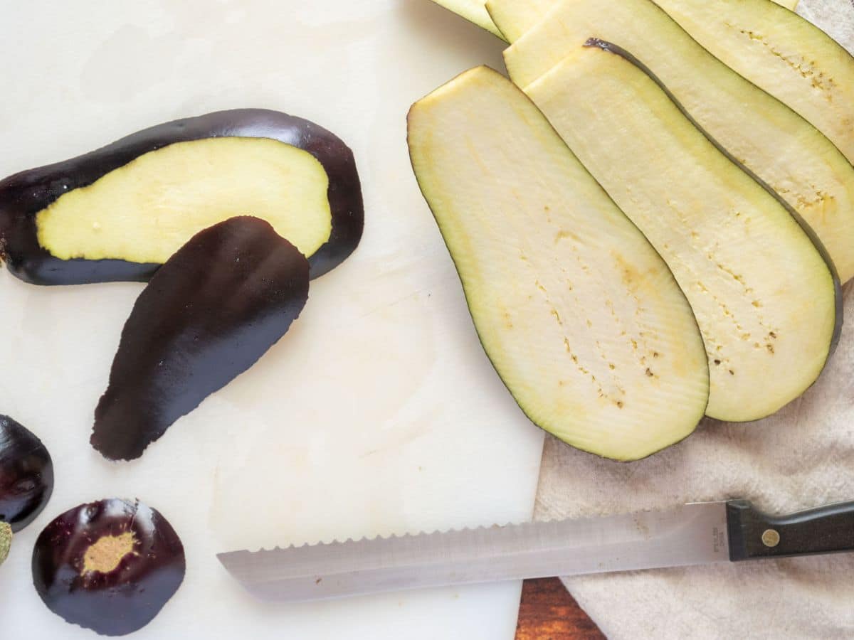 Thin slices of eggplant and eggplant peels are on a cutting board next to a serrated knife.