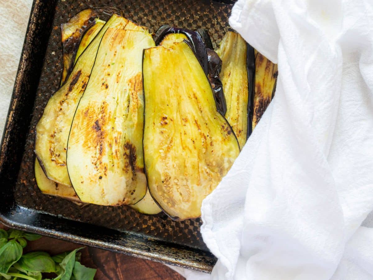Sliced, roasted eggplant in a dark baking tray partially covered by a white cloth.