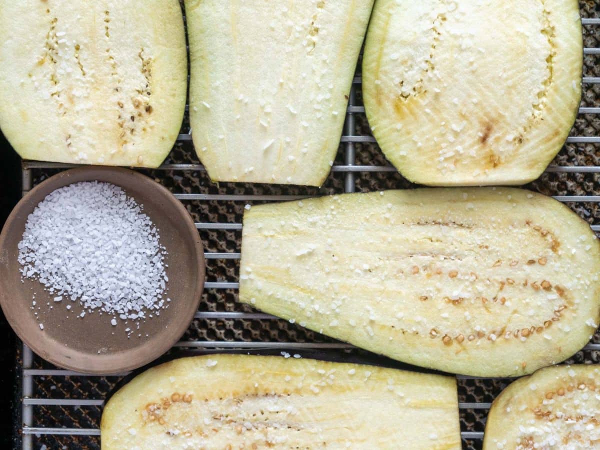 Sliced raw eggplant sprinkled with salt on a wire rack, next to a small bowl of coarse salt.