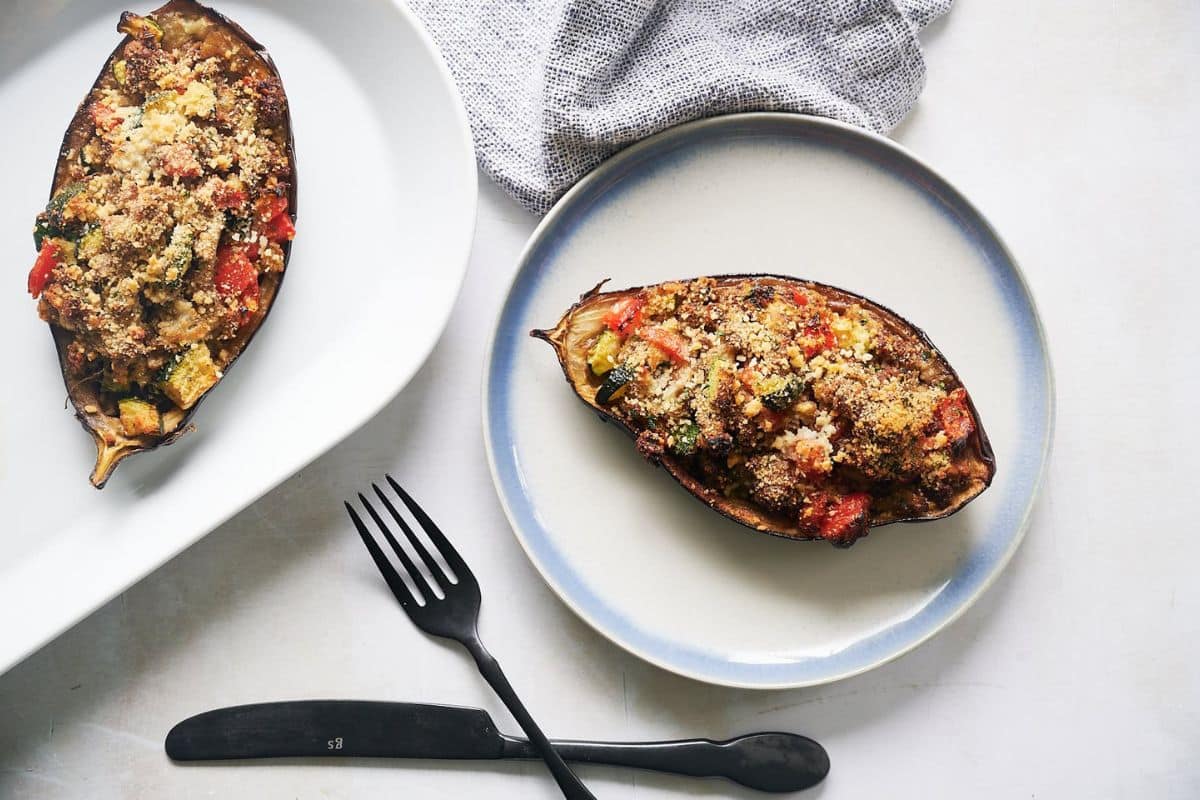 A plate with a stuffed and baked eggplant half, filled with vegetables and topped with breadcrumbs-one of the delicious ways to cook eggplant. A serving platter with another stuffed eggplant and cutlery are beside the plate.