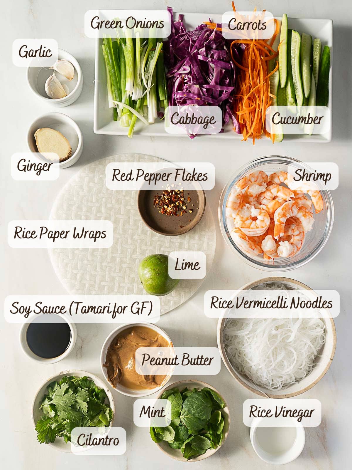A delicious selection of fresh and prepared ingredients for shrimp summer rolls, including vegetables, herbs, rice paper wraps, rice noodles, and dipping sauces, beautifully arranged on a white surface.