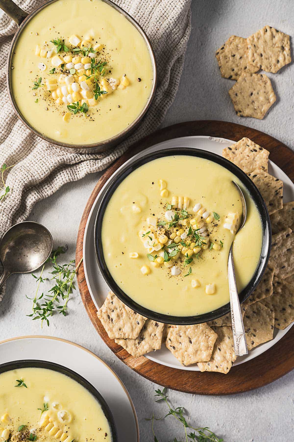Three bowls of creamy corn soup garnished with corn kernels and herbs, served with crackers on a plate; a spoon and napkin are nearby.