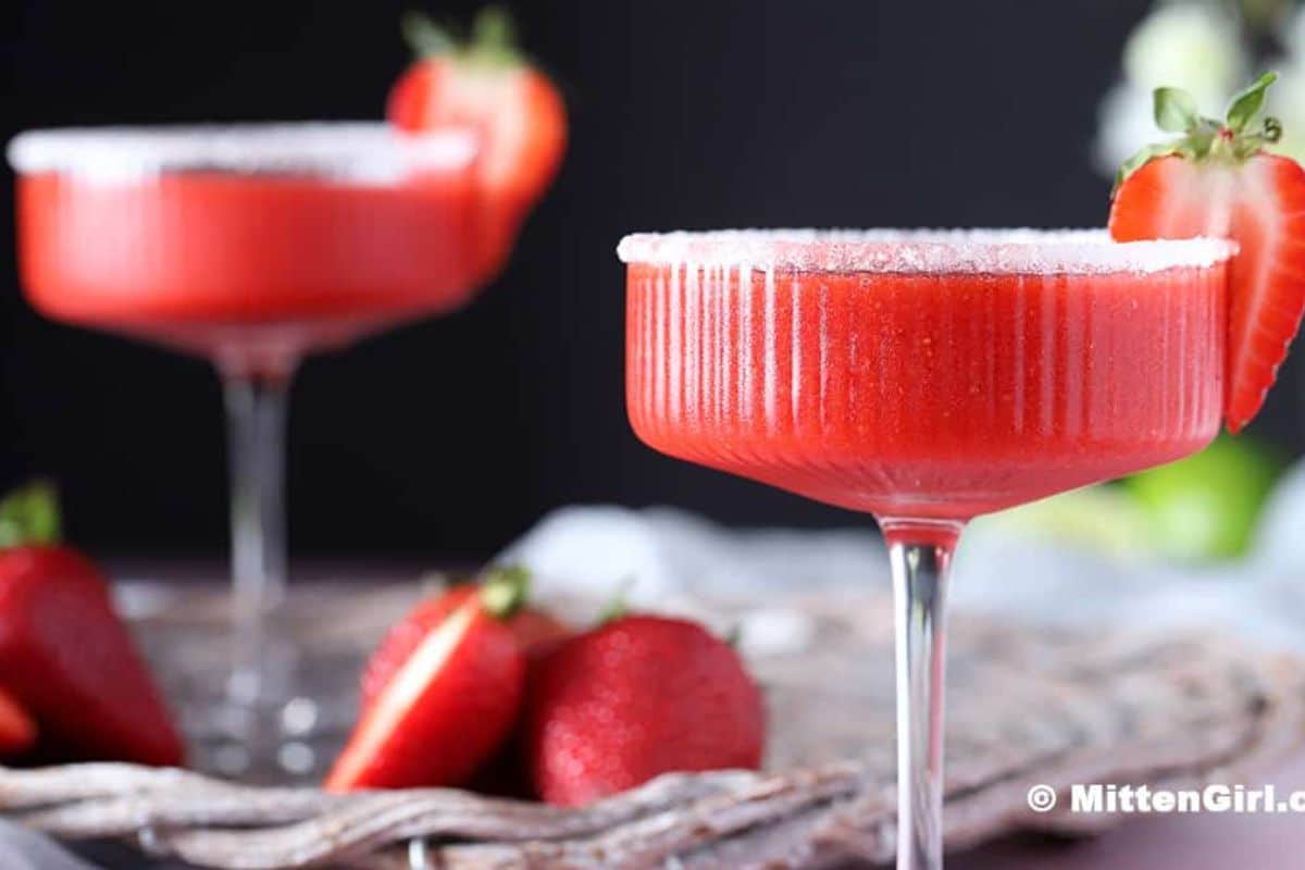 Two glasses of refreshing, non alcoholic frozen strawberry margarita with sugar rims, garnished with fresh strawberries; whole and sliced strawberries placed nearby-perfect for summer drinks.