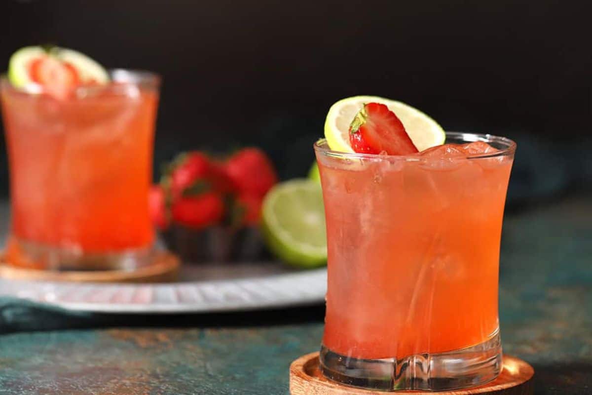 Two glasses of a refreshing red iced drink garnished with lime and strawberry slices, perfect for summer drinks. Fresh strawberries and a lime sit in the background, making this a delicious non alcoholic treat.