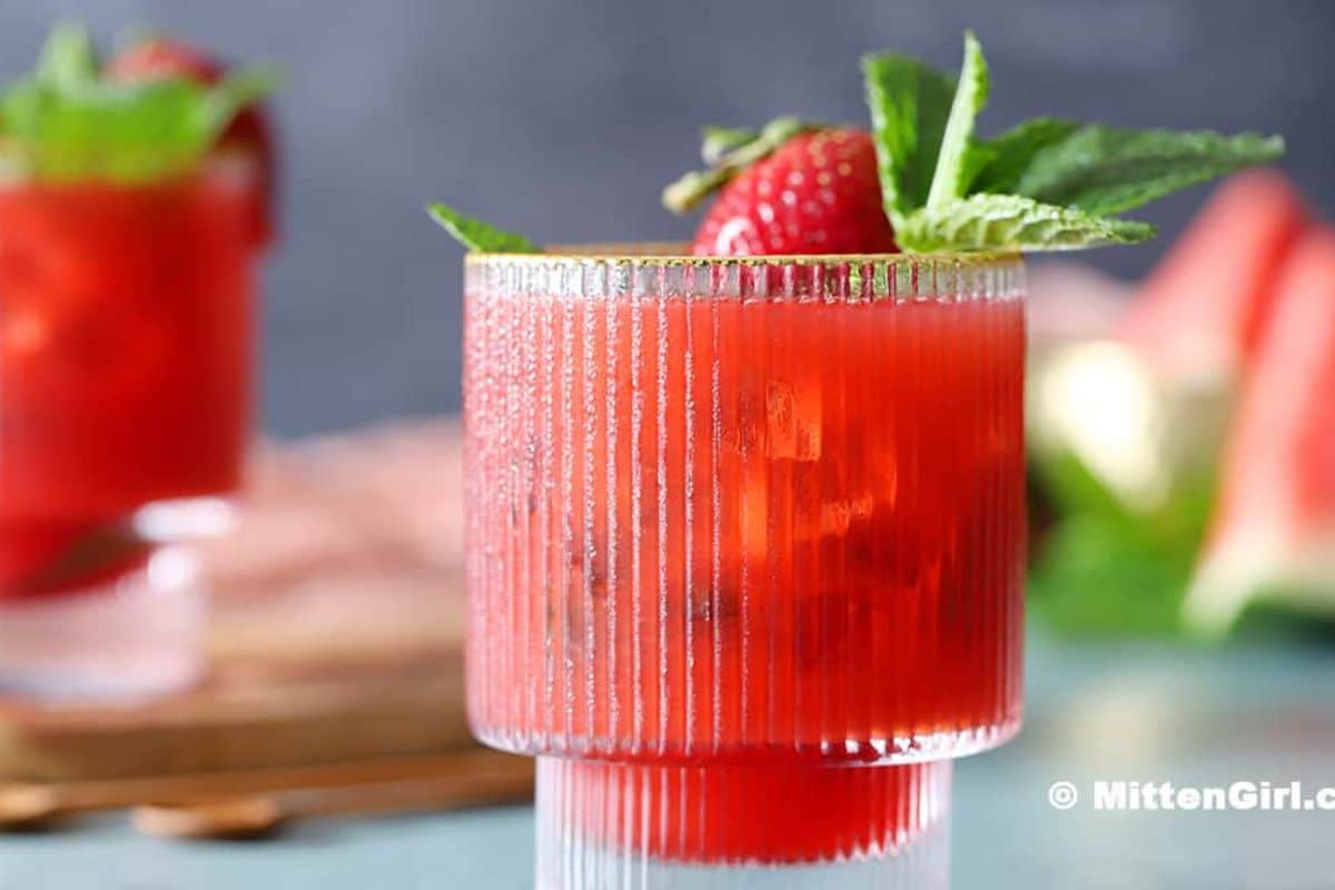 A glass filled with a refreshing red beverage, garnished with a strawberry and mint leaves, sits on a table with another summer drink and slices of watermelon in the blurred background.