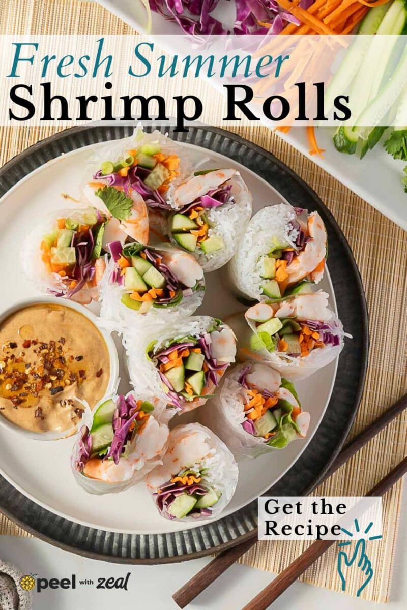 A plate of shrimp summer rolls filled with crisp vegetables and served with peanut dipping sauce, surrounded by fresh, colorful ingredients.
