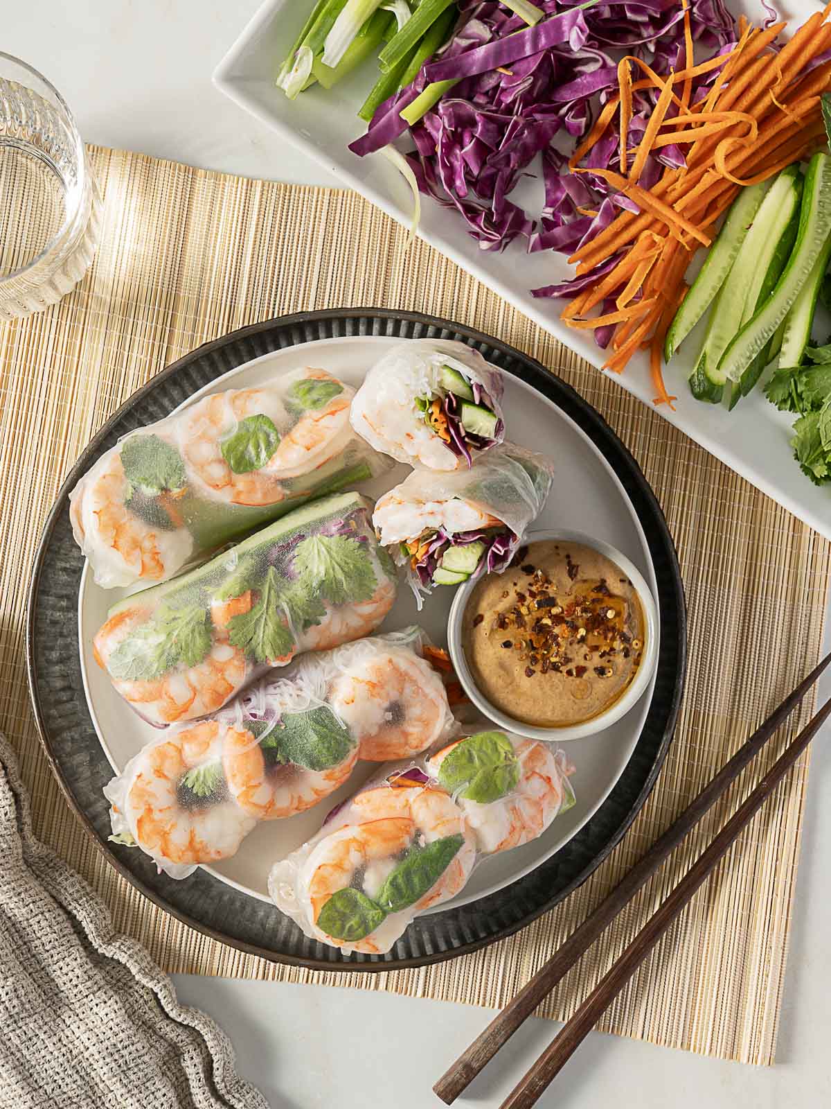 Plate of shrimp spring rolls with fresh herbs and vegetables, served with dipping sauce; additional sliced vegetables on a separate tray nearby; chopsticks and a glass of water beside the plate.