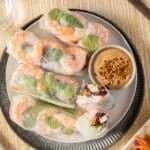 A plate of shrimp spring rolls with fresh vegetables, served with a small bowl of peanut dipping sauce on the side.