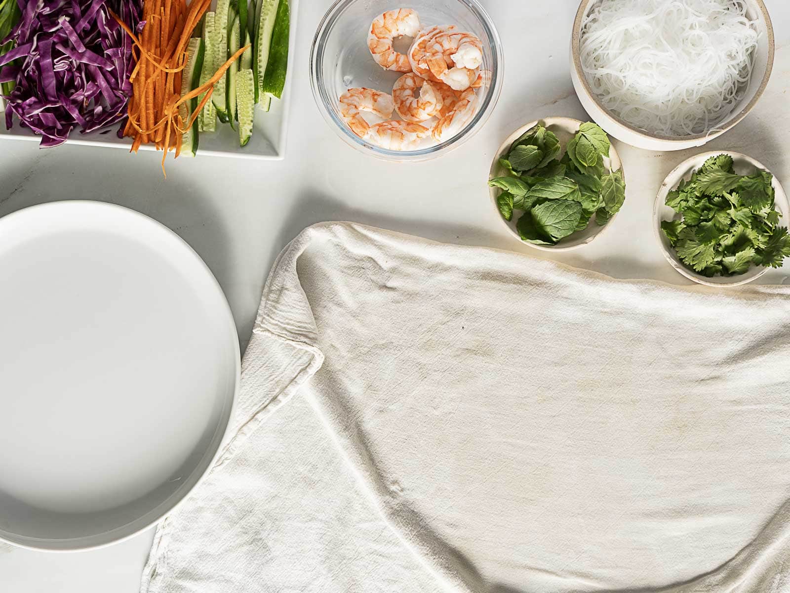 Ingredients for making spring rolls, including shrimp, rice noodles, mint, cilantro, shredded carrots, cucumbers, red cabbage, and an empty plate, arranged on a white surface.