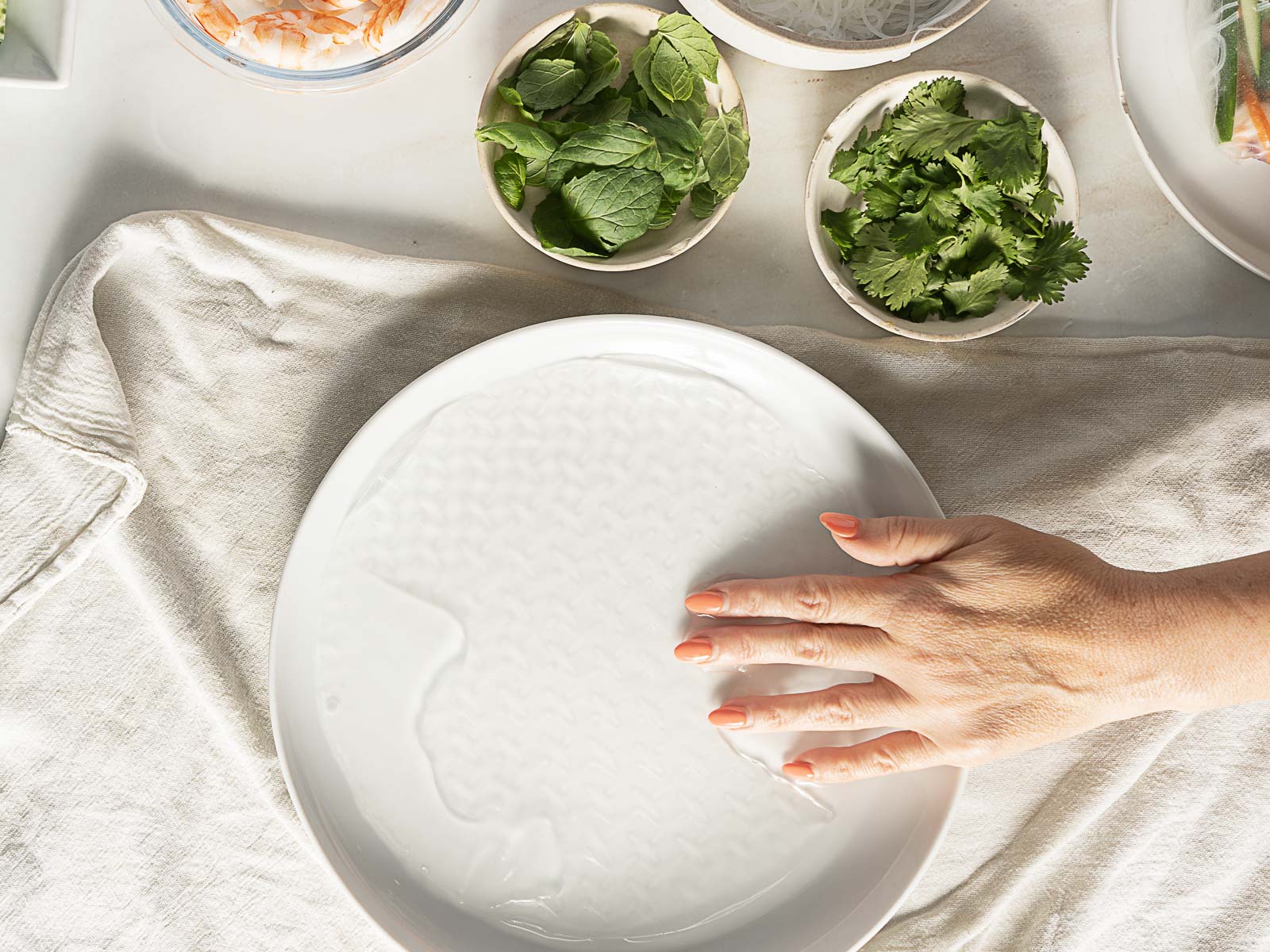 A hand presses a sheet of rice paper onto a plate of water, surrounded by bowls of fresh herbs and vegetables on a cloth.