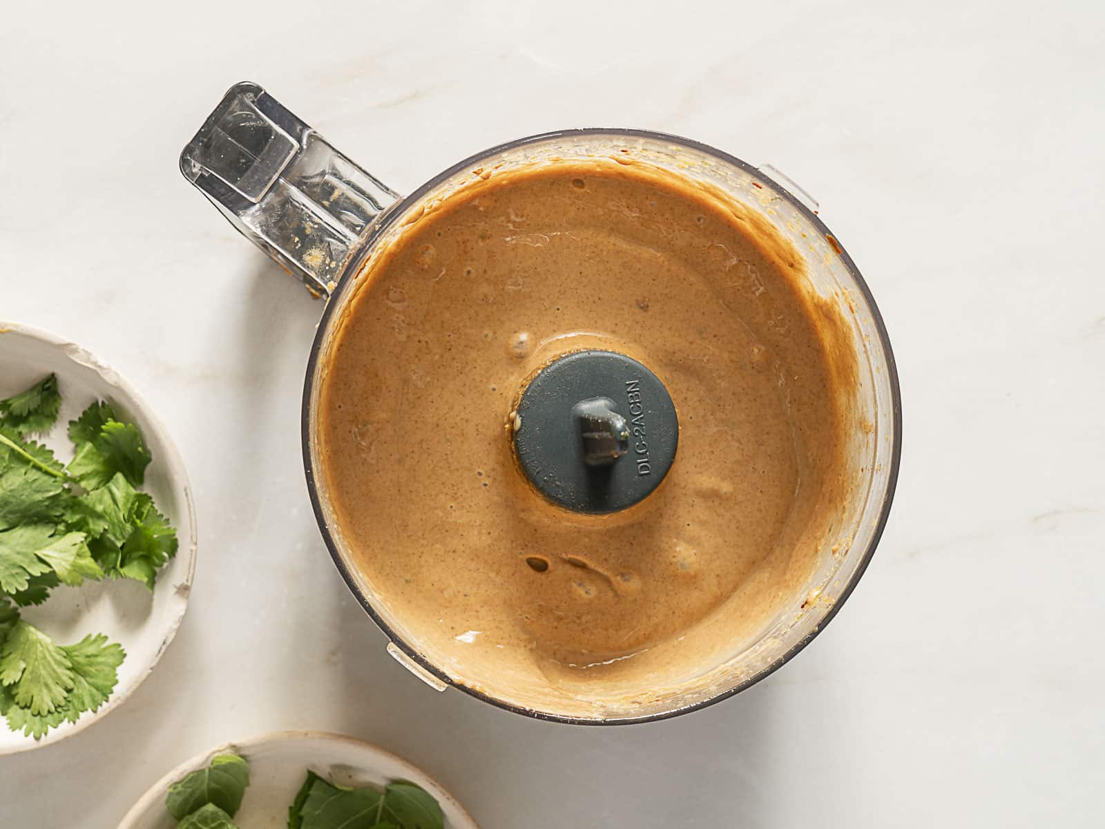 Top view of a blender containing a smooth, brown sauce; fresh cilantro leaves are in small bowls nearby on a light surface.