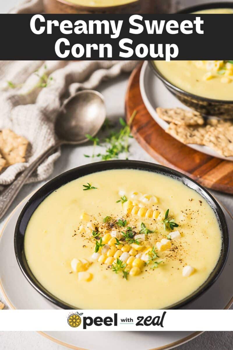 A bowl of creamy corn bisque garnished with corn kernels, black pepper, and fresh herbs, served with crackers and a spoon on the side.