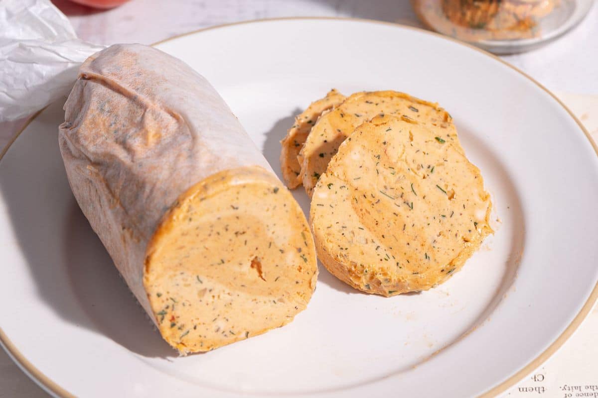 A roll of herbed compound butter, perfect for summer recipes, is partially wrapped in parchment, with two round slices cut and placed on a white plate.