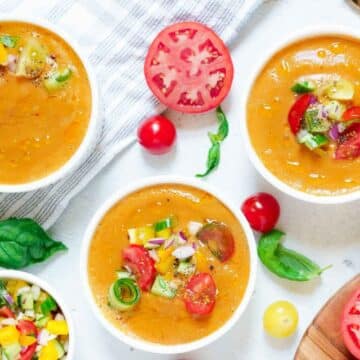 Three bowls of gazpacho, a classic fresh summer soup, garnished with chopped vegetables and herbs, are surrounded by fresh tomato slices, tomatoes, basil leaves, and a spoon on a white surface.