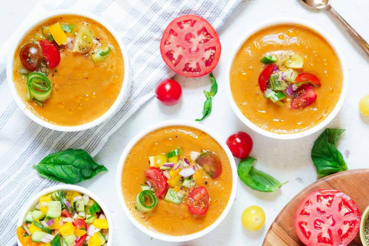 Three bowls of gazpacho, a classic fresh summer soup, garnished with chopped vegetables and herbs, are surrounded by fresh tomato slices, tomatoes, basil leaves, and a spoon on a white surface.