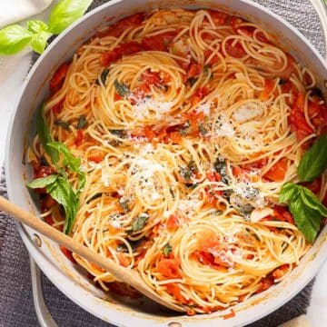 A pot of spaghetti with tomato sauce, basil leaves, and grated cheese, with a wooden spoon resting inside; cherry tomatoes and cheese nearby make this one of the best summer recipes using ripe tomatoes.
