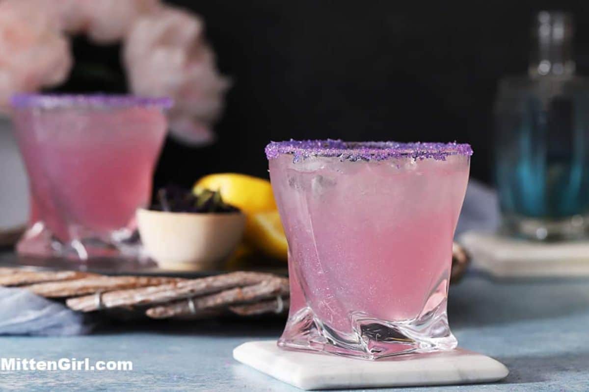 Two glasses of pink Refreshing Summer Drinks with purple sugar rims sit on marble coasters, accompanied by a lemon, a small bowl, and a bottle in the background.