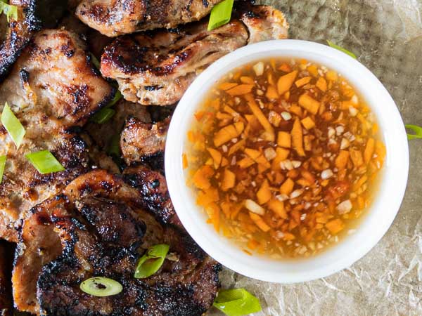 Grilled meat slices garnished with green onions are served next to a small bowl of dipping sauce containing chopped garlic and orange vegetables.