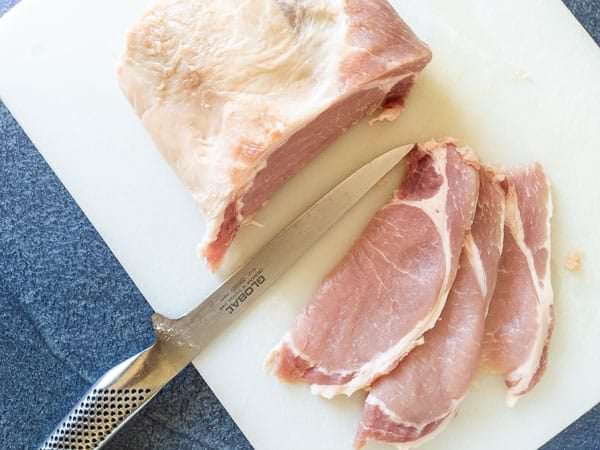 A partially sliced raw pork loin with three slices and a chef's knife on a white cutting board.