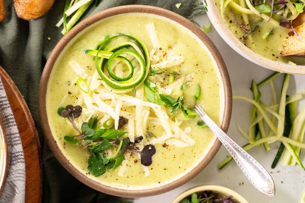 A bowl of creamy zucchini soup, a fresh summer soup topped with shredded cheese, microgreens, spiralized zucchini, and cracked black pepper, with a spoon resting inside.