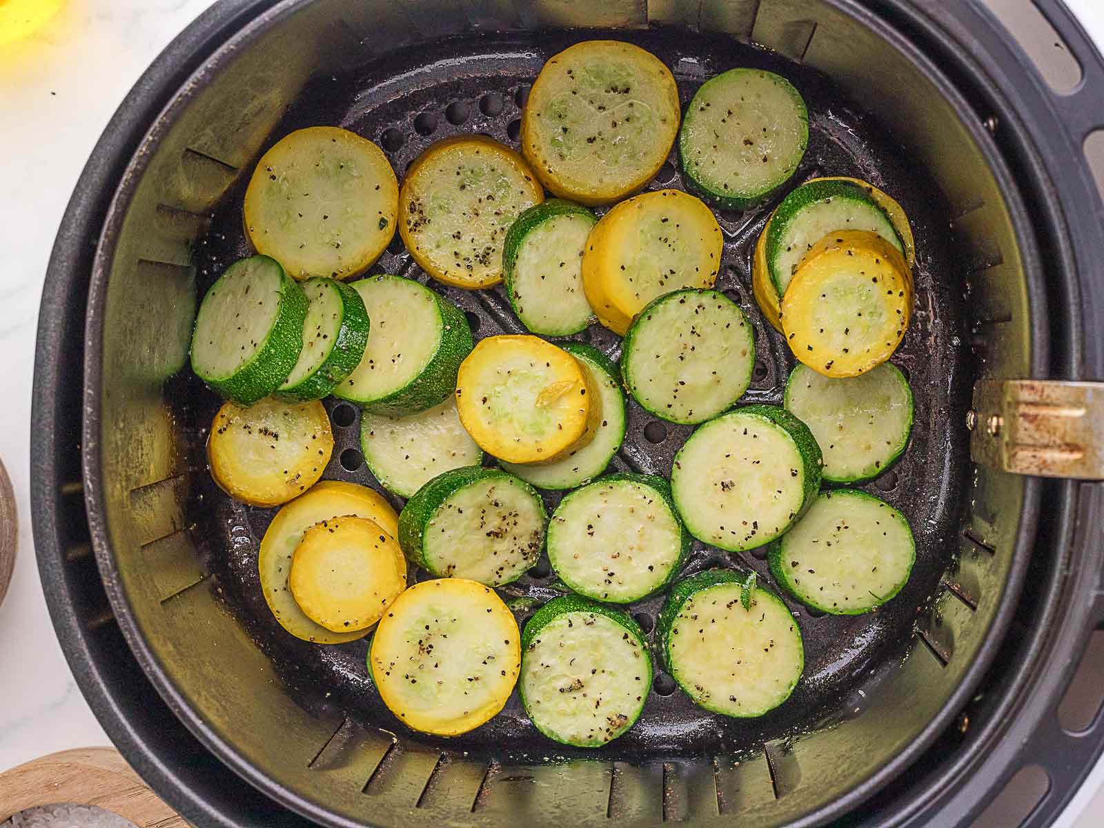 Sliced yellow and green squash seasoned with pepper are arranged in the basket of an air fryer, ready to be cooked.