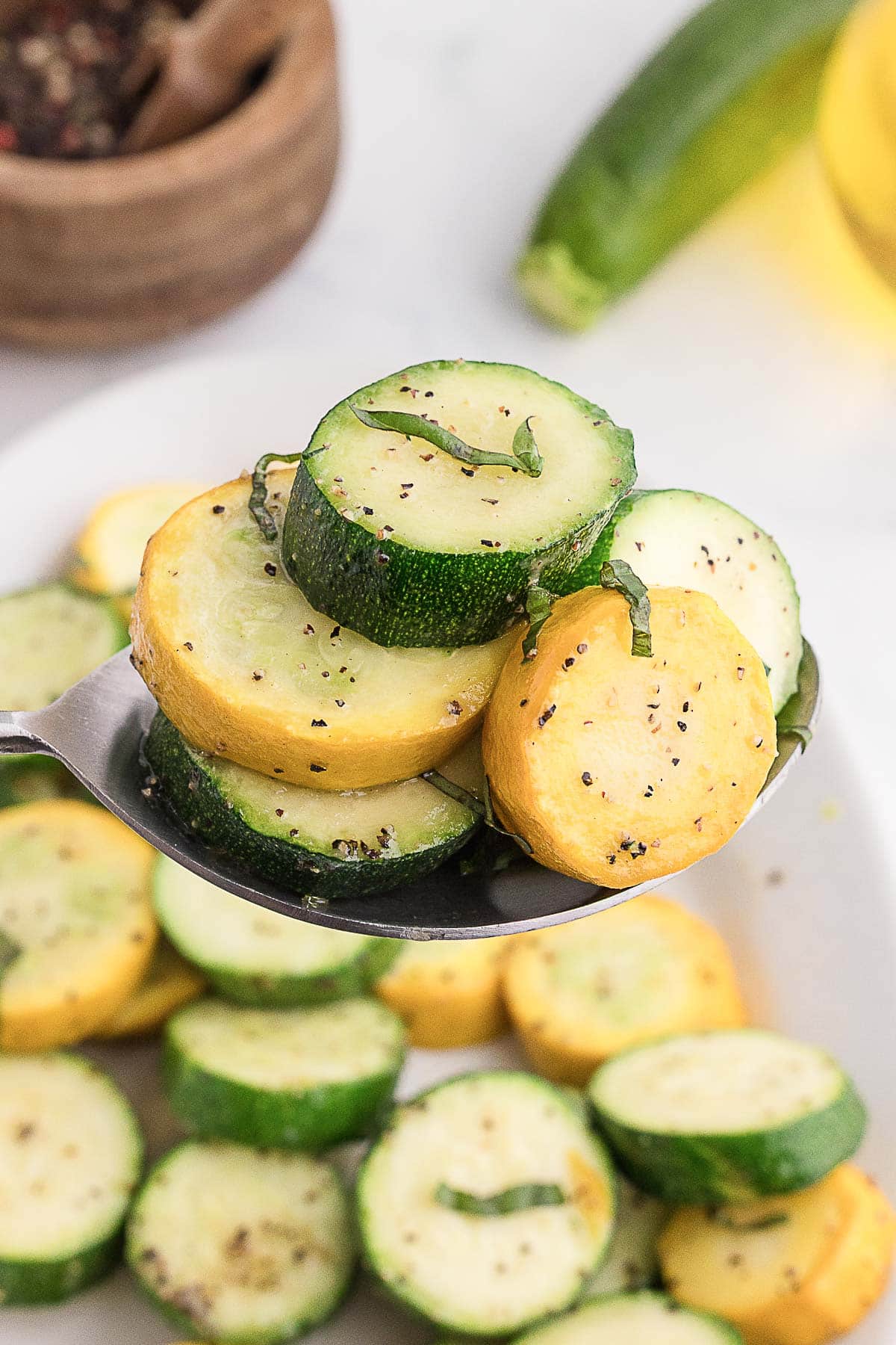 A close-up of a spoon holding slices of cooked yellow squash and zucchini, seasoned with pepper and garnished with herbs.