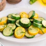 A white plate with sliced, cooked yellow squash and zucchini, seasoned with black pepper and garnished with fresh herbs.