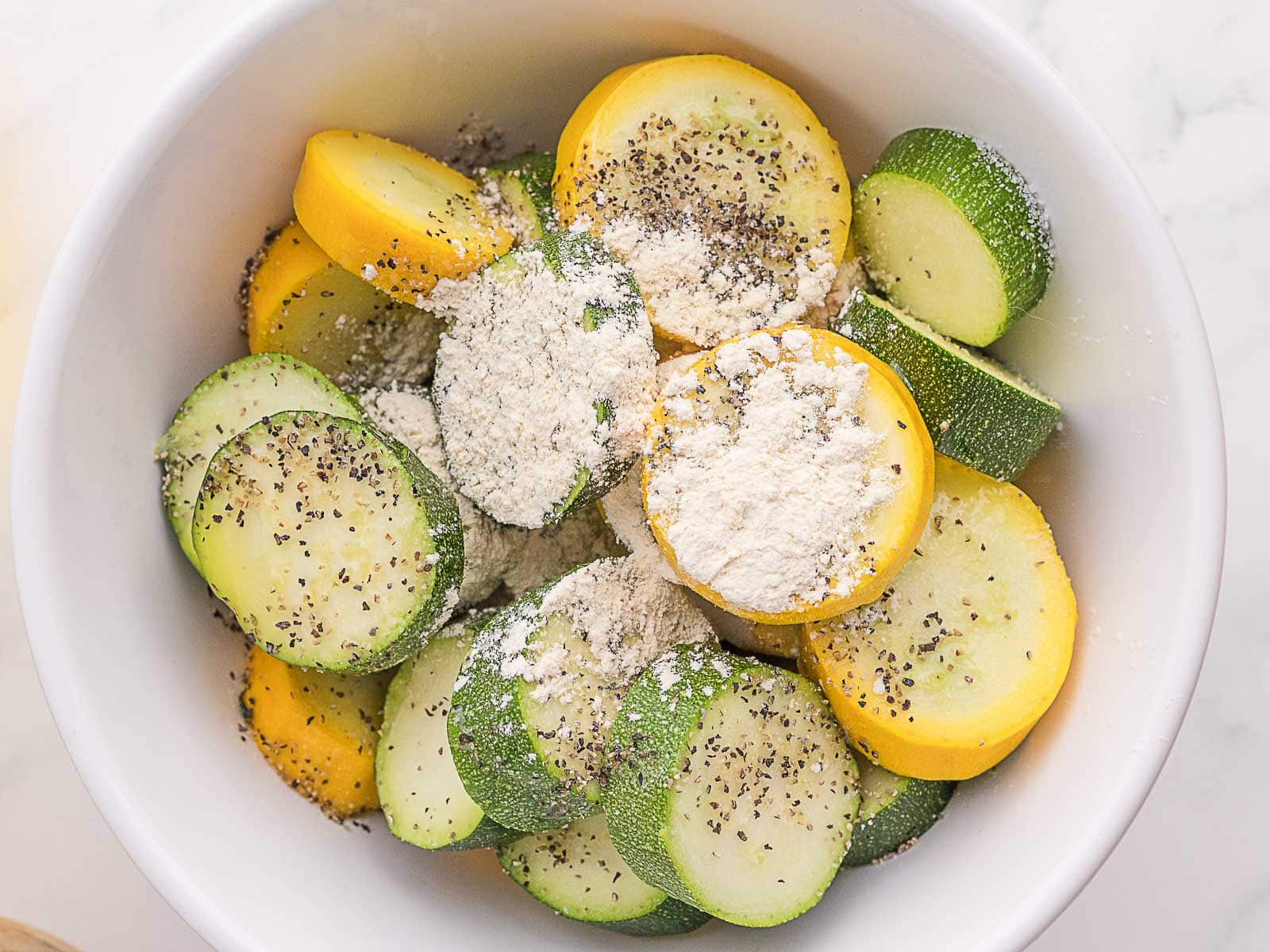 A white bowl filled with sliced yellow and green zucchini topped with flour and black pepper.
