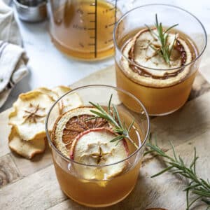 Two glasses of amber-colored beverage garnished with dried apple slices, citrus, and rosemary sprigs on a wooden surface with a jar of liquid and more dried fruit nearby.
