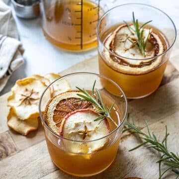 Two glasses of amber-colored beverage garnished with dried apple slices, citrus, and rosemary sprigs on a wooden surface with a jar of liquid and more dried fruit nearby.