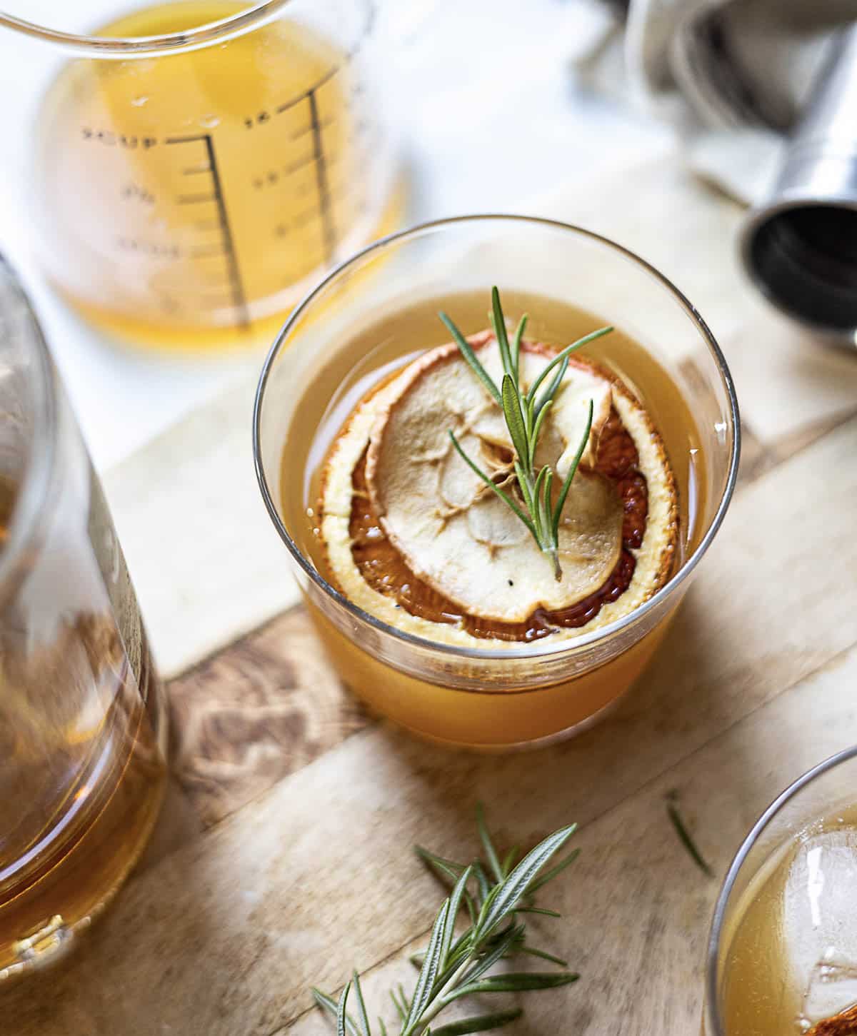 A glass of amber-colored drink garnished with a dried apple slice and rosemary sits on a wooden surface next to a measuring cup and cocktail tools.