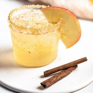 A glass of apple cocktail with a sugar rim and apple slice garnish sits on a white plate beside two cinnamon sticks.
