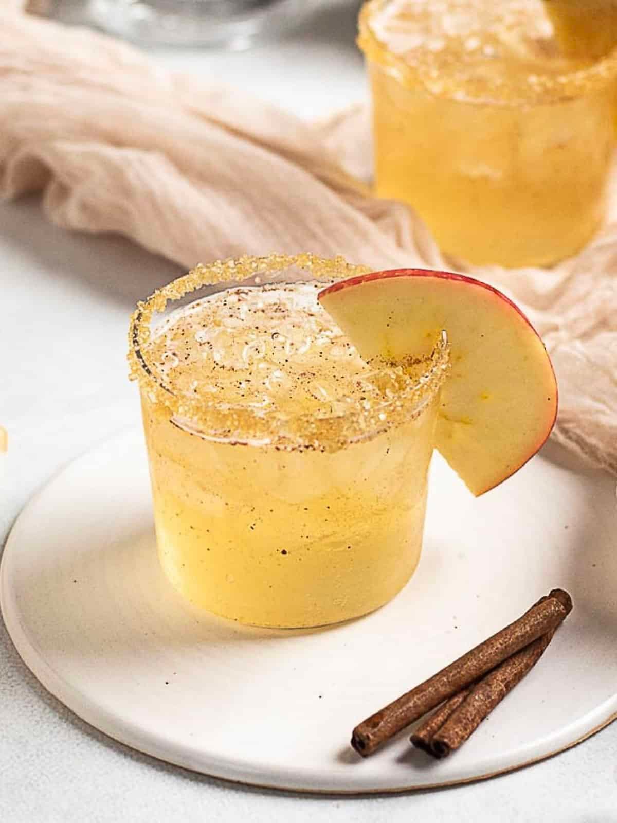 A glass of yellow cocktail with a sugared rim and apple slice garnish sits on a white plate beside two cinnamon sticks.