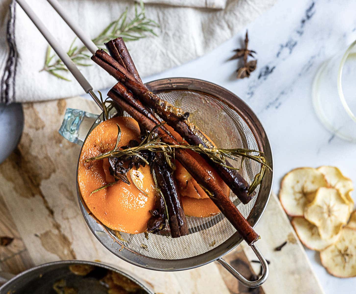 A metal strainer holds cooked apple slices, cinnamon sticks, rosemary, and star anise, set on a wooden surface beside dried apple slices and a glass.