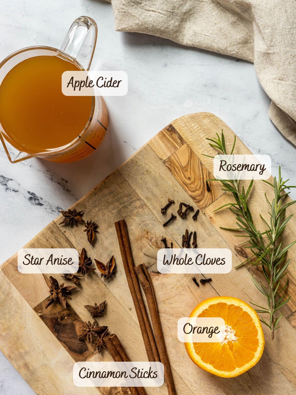 A wooden board with whole cloves, rosemary, orange, star anise, and cinnamon sticks sits next to a measuring cup of apple cider syrup on a marble surface.