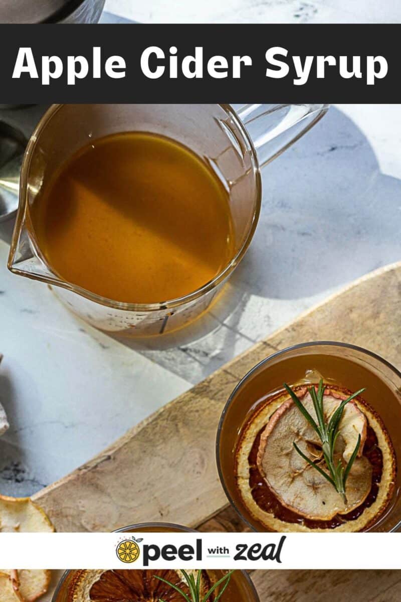 A glass measuring cup filled with cider syrup sits next to a drink garnished with dried apple slices and rosemary on a wooden board.