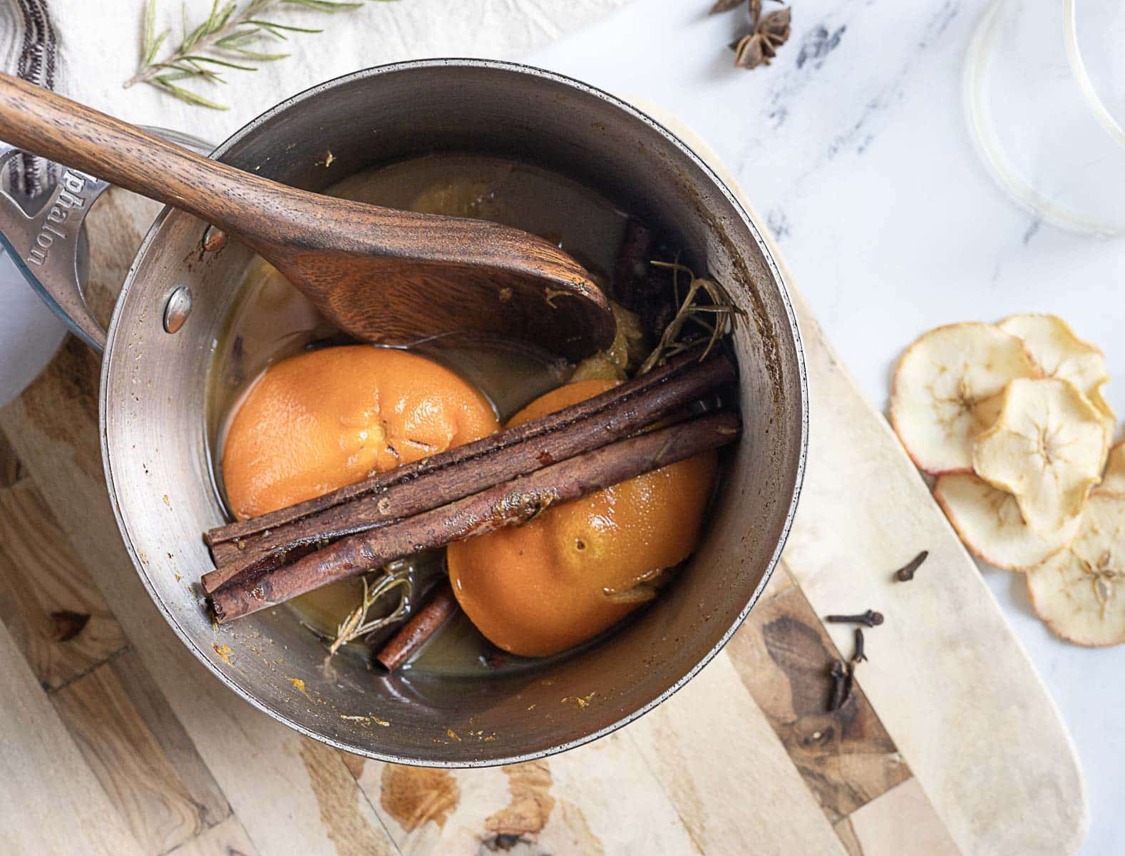 A saucepan contains orange halves, cinnamon sticks, and herbs, with a wooden spoon resting inside. Dried apple slices and whole cloves are on the table nearby.