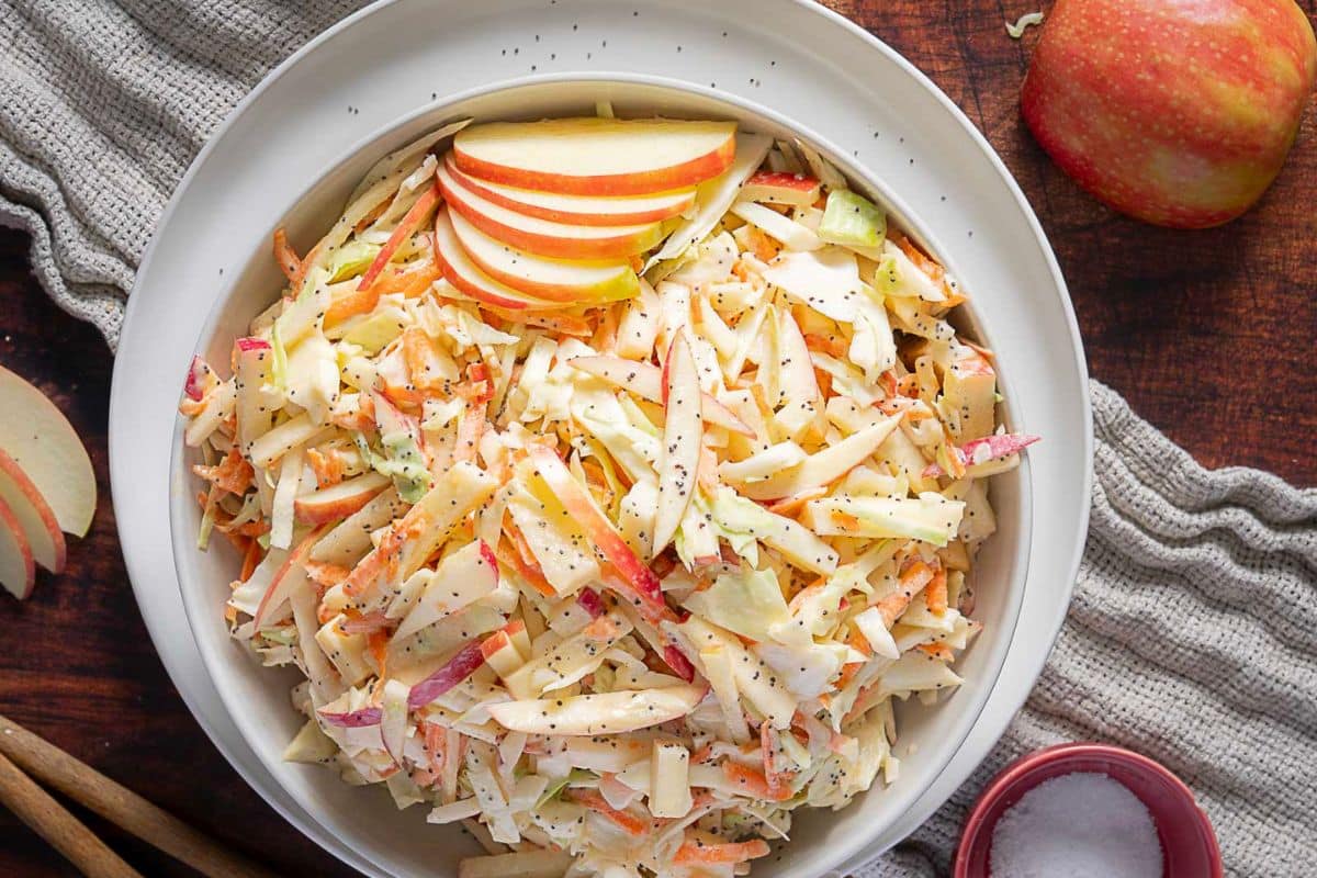 A bowl of apple coleslaw topped with sliced apples, with an apple, a napkin, and a small bowl of salt on a wooden surface-perfect for easy side dishes at dinner or quick weeknight sides.