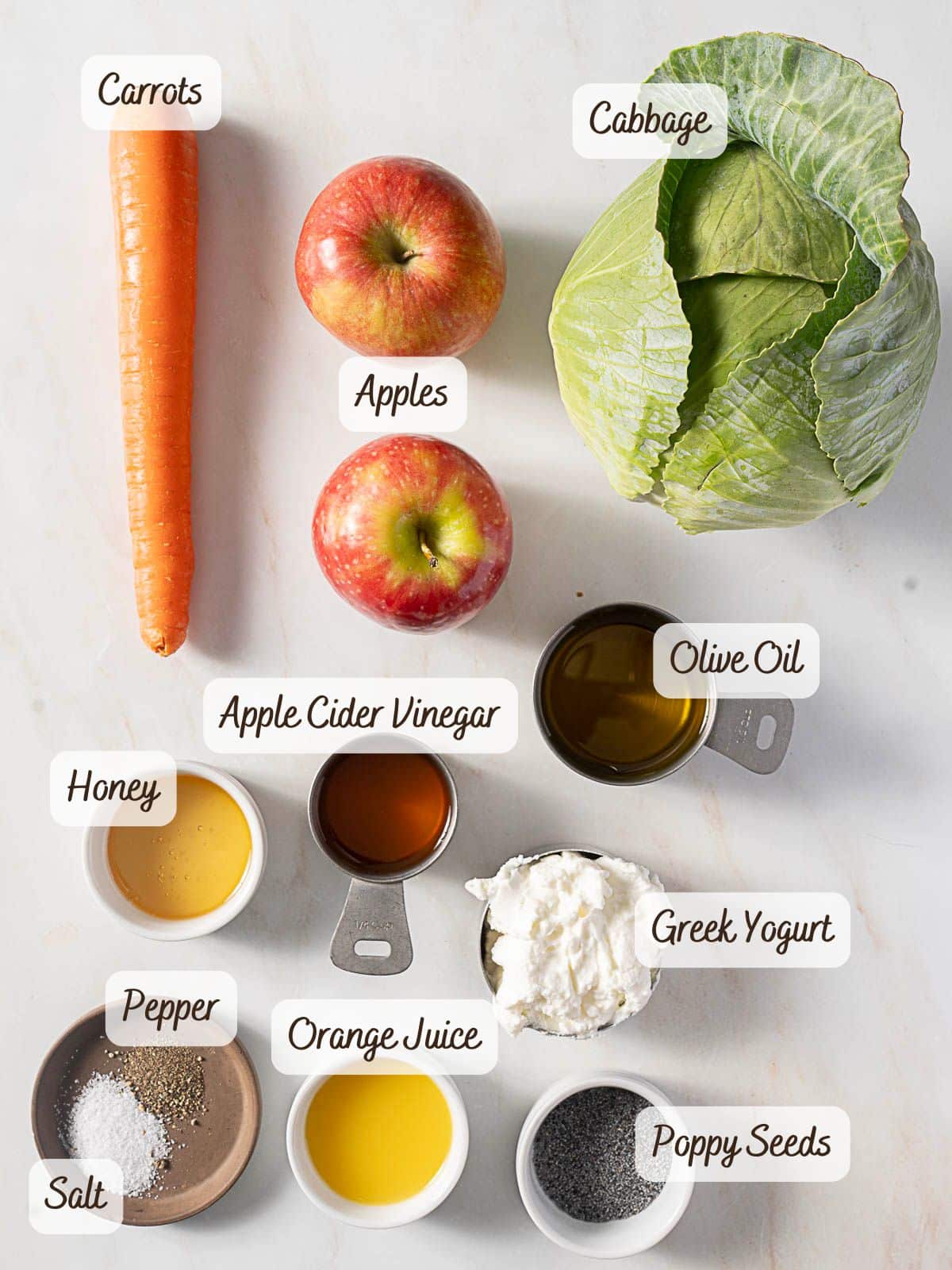 Overhead view of ingredients for a salad, including carrot, cabbage, apples, honey, apple cider vinegar, olive oil, Greek yogurt, orange juice, poppy seeds, salt, and pepper.