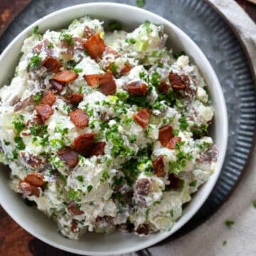 A bowl of potato salad topped with chopped bacon and fresh herbs, placed on a metal plate with a napkin and wooden spoon nearby—one of the best side dishes sure to be a show-stealing side at any gathering.