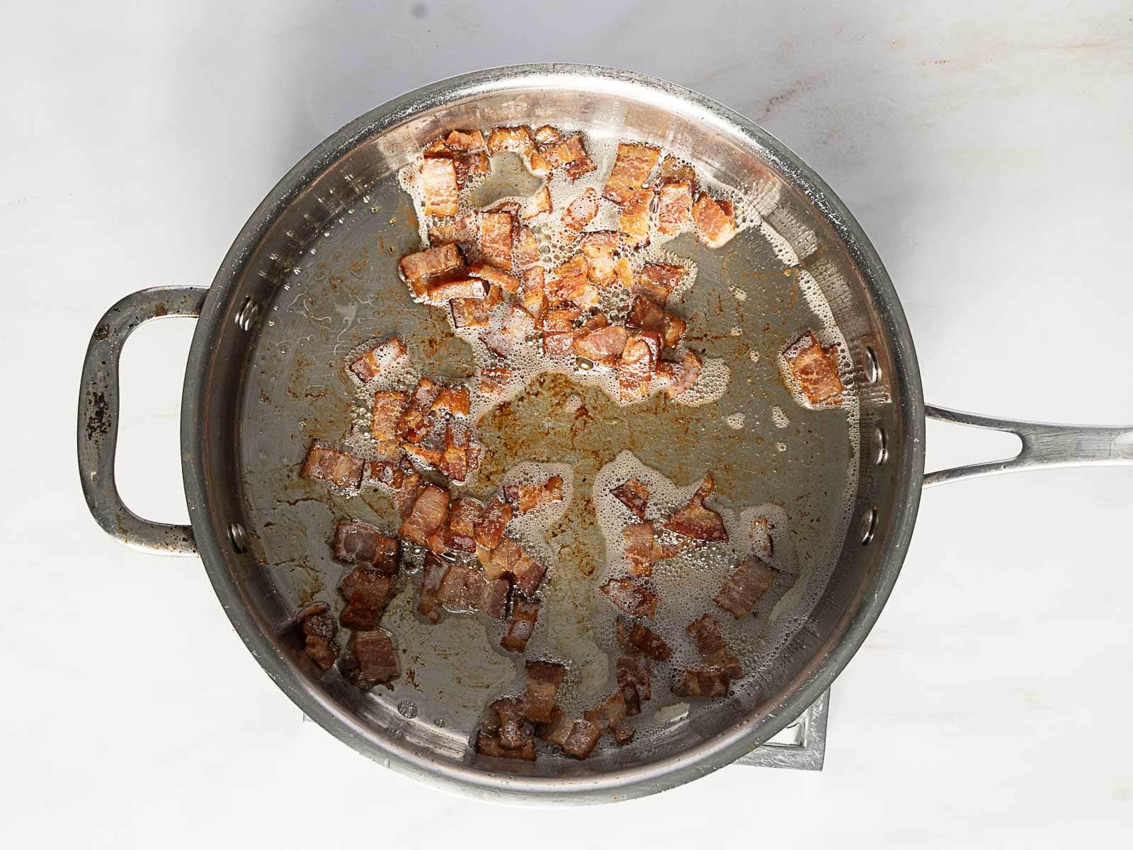 A stainless steel pan with pieces of cooked, browned bacon and some rendered fat on a white surface.