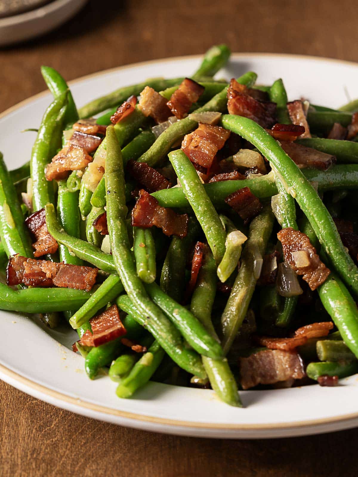 A plate of cooked green beans mixed with pieces of bacon and diced onions on a white dish.