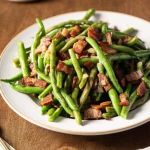 A plate of cooked green beans mixed with chopped bacon pieces on a wooden table.