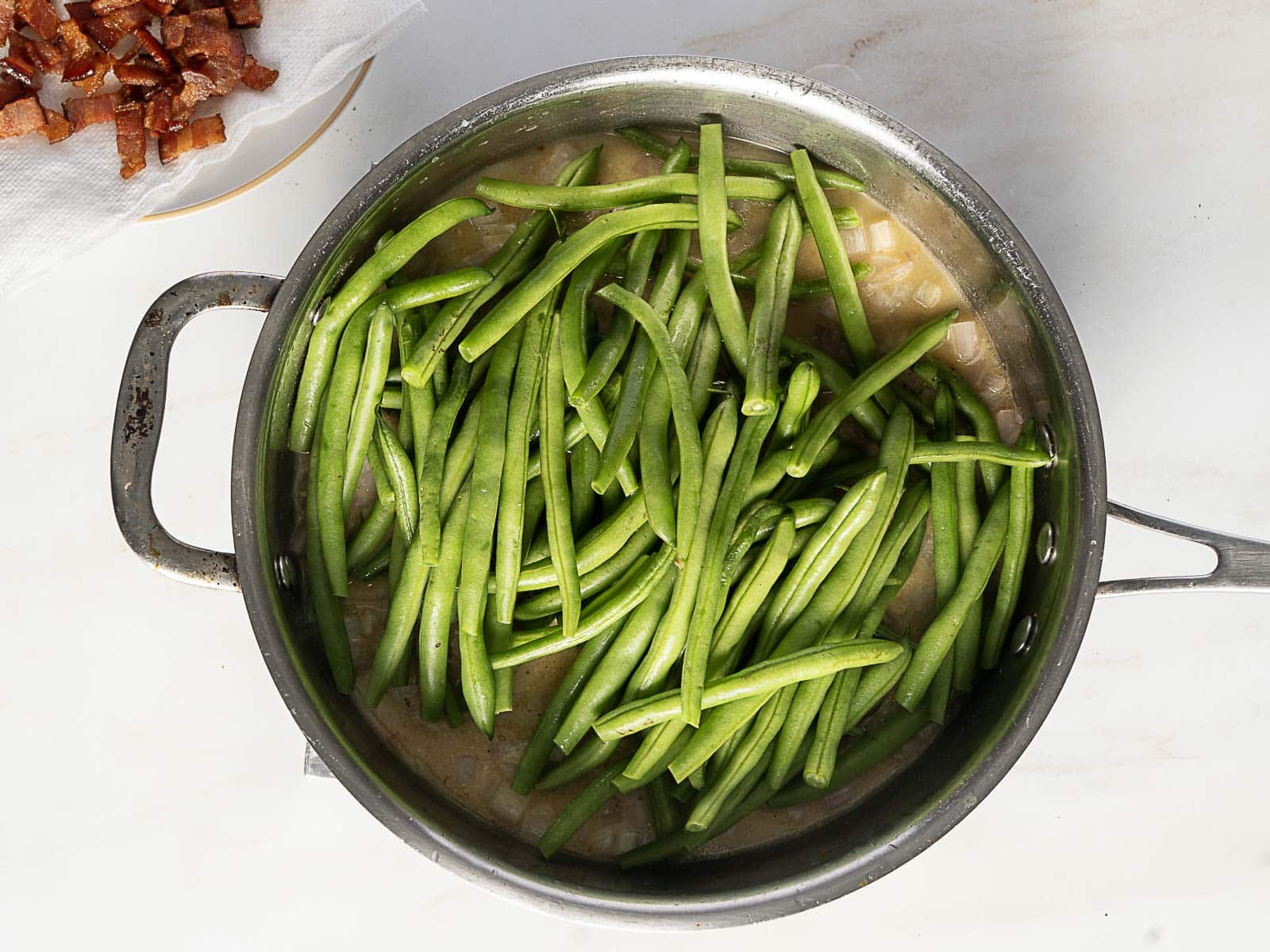 A stainless steel pot filled with uncooked green beans in a light sauce on a white surface, with chopped bacon on a plate nearby.