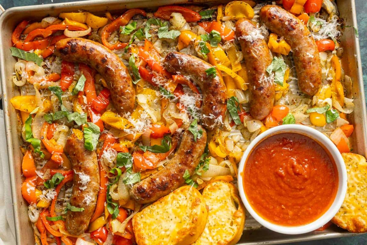 Perfect for busy School Nights, this sheet pan features roasted sausages, sliced bell peppers, onions, fresh basil, grated cheese, toasted bread, and a bowl of marinara sauce.