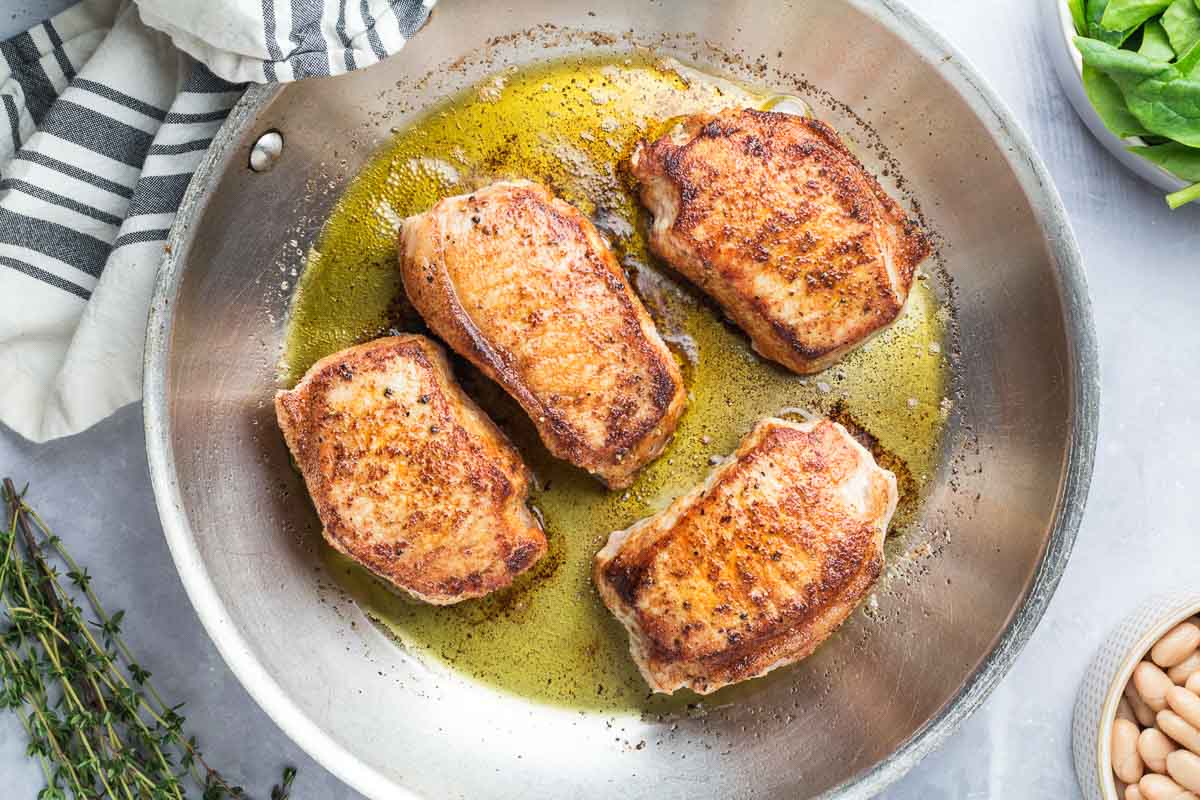 Skillet Pork Chops are searing in a stainless steel skillet with oil-perfect for busy school nights. Fresh thyme, a towel, and a bowl of spinach sit nearby, ready to complete this quick and delicious meal.