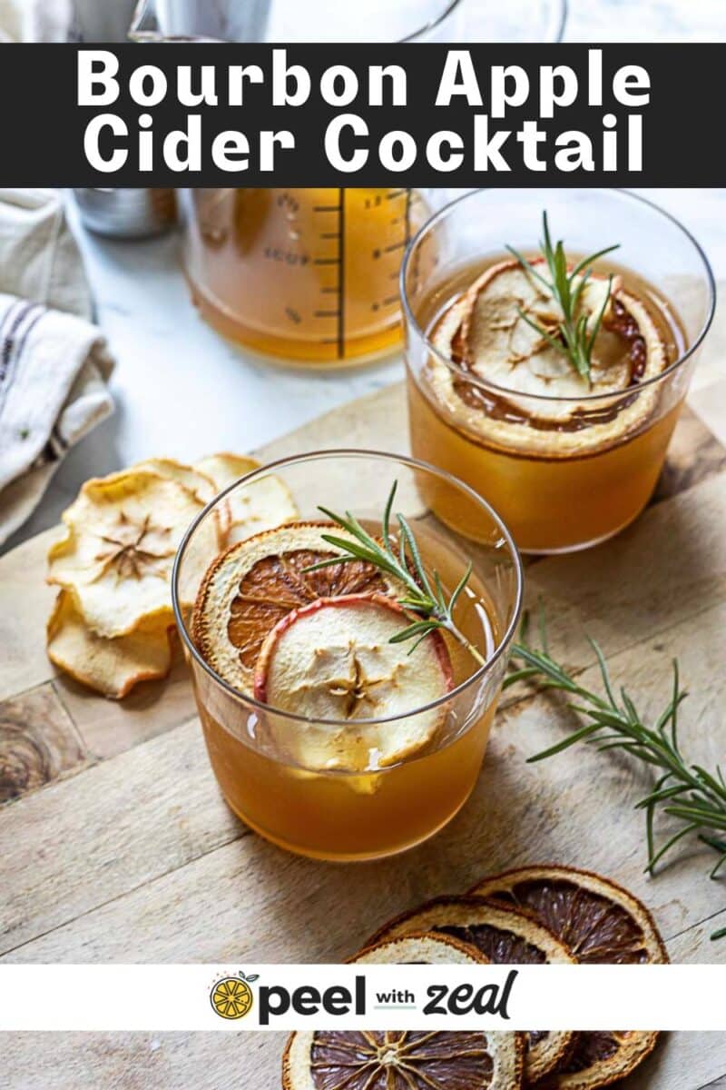Two glasses of bourbon apple cider cocktail garnished with dried apple slices and rosemary rest on a wooden surface, surrounded by dried fruit slices.