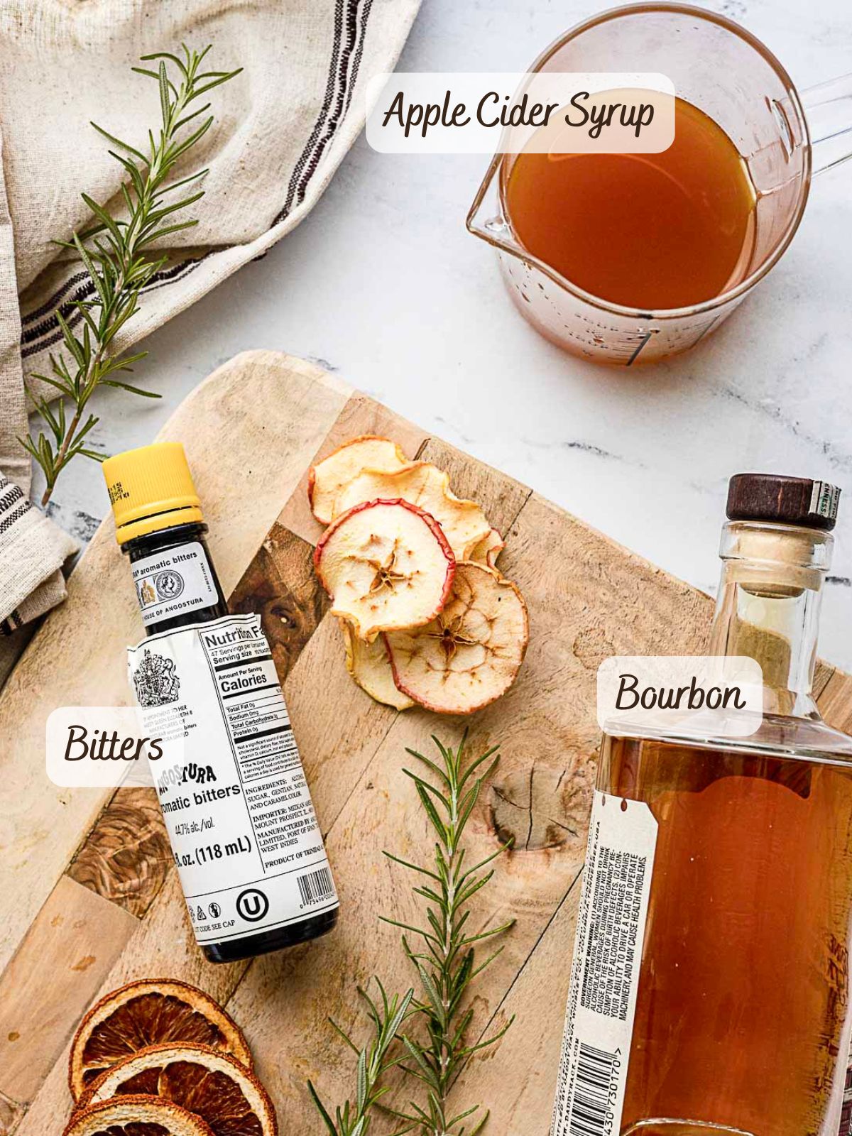 A bottle of bitters, bourbon, a measuring cup of apple cider syrup, dried apple slices, and rosemary on a wooden cutting board-perfect ingredients for crafting a bourbon apple cider cocktail.