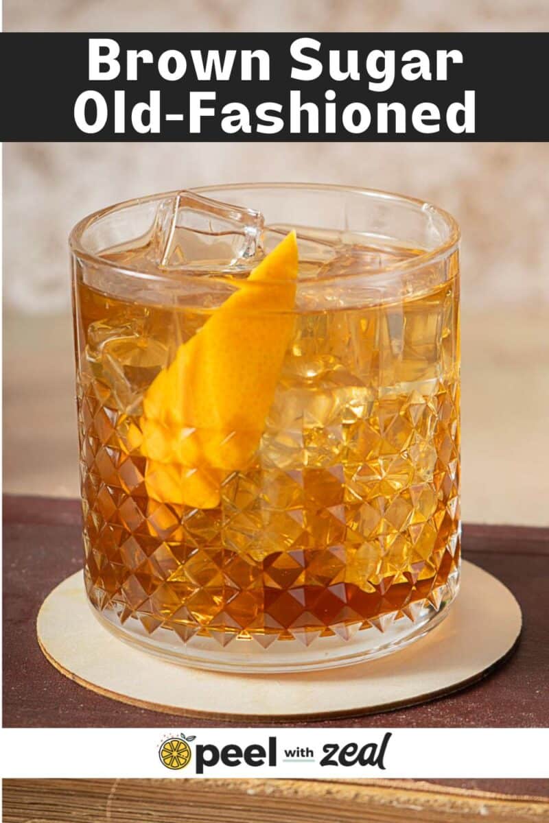 A glass of scotch old fashioned with brown sugar, ice, and an orange peel garnish sits on a coaster.